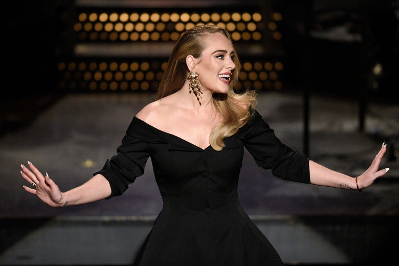 Adele in a black dress smiles and extends her arms out to her sides onstage