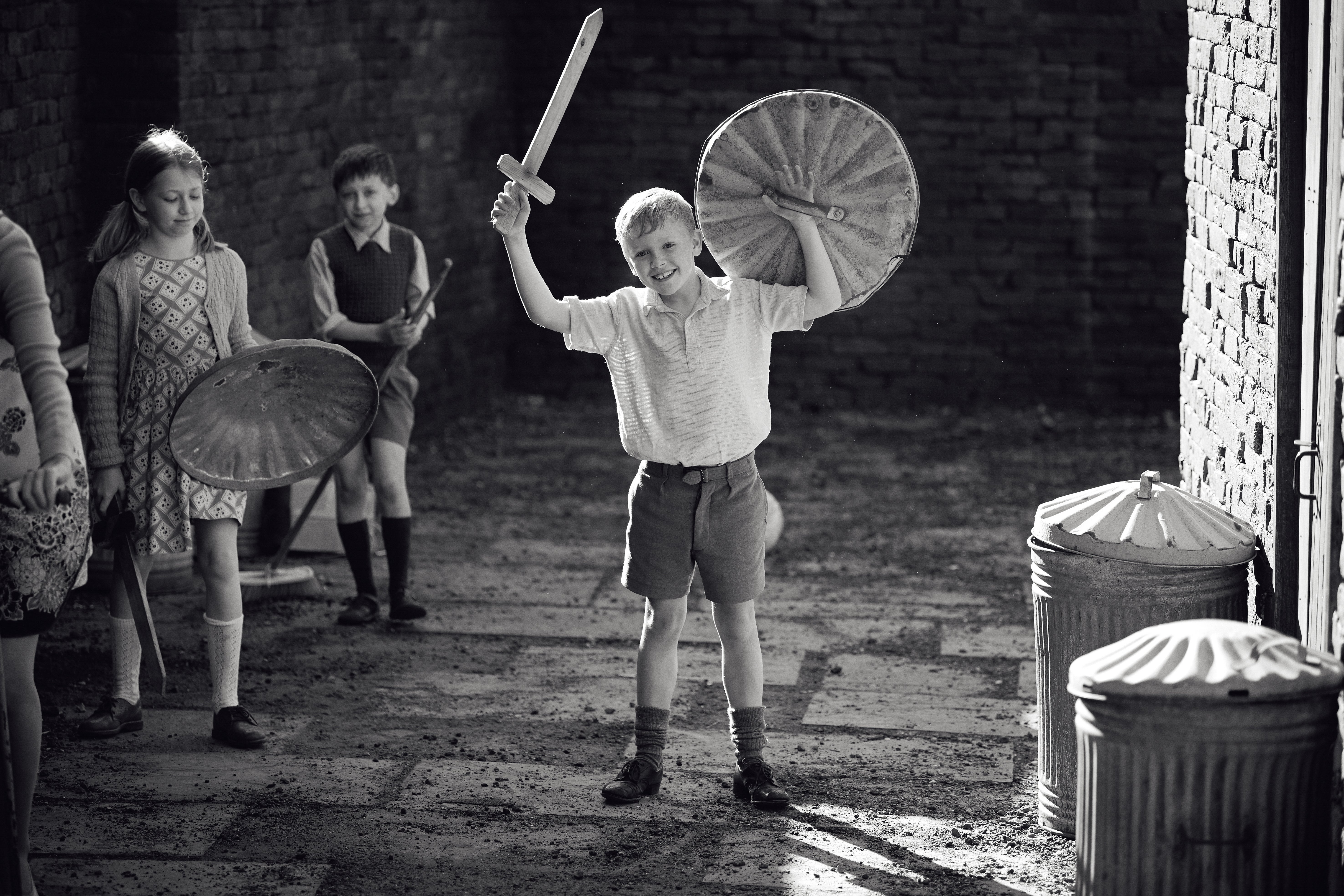 A black-and-white still of a smiling, doe-eyed blond child in short shorts and long socks holding up a toy sword and the lid of a trash can like a shield. Other kids stand nearby also holding toy swords and trash can lids.
