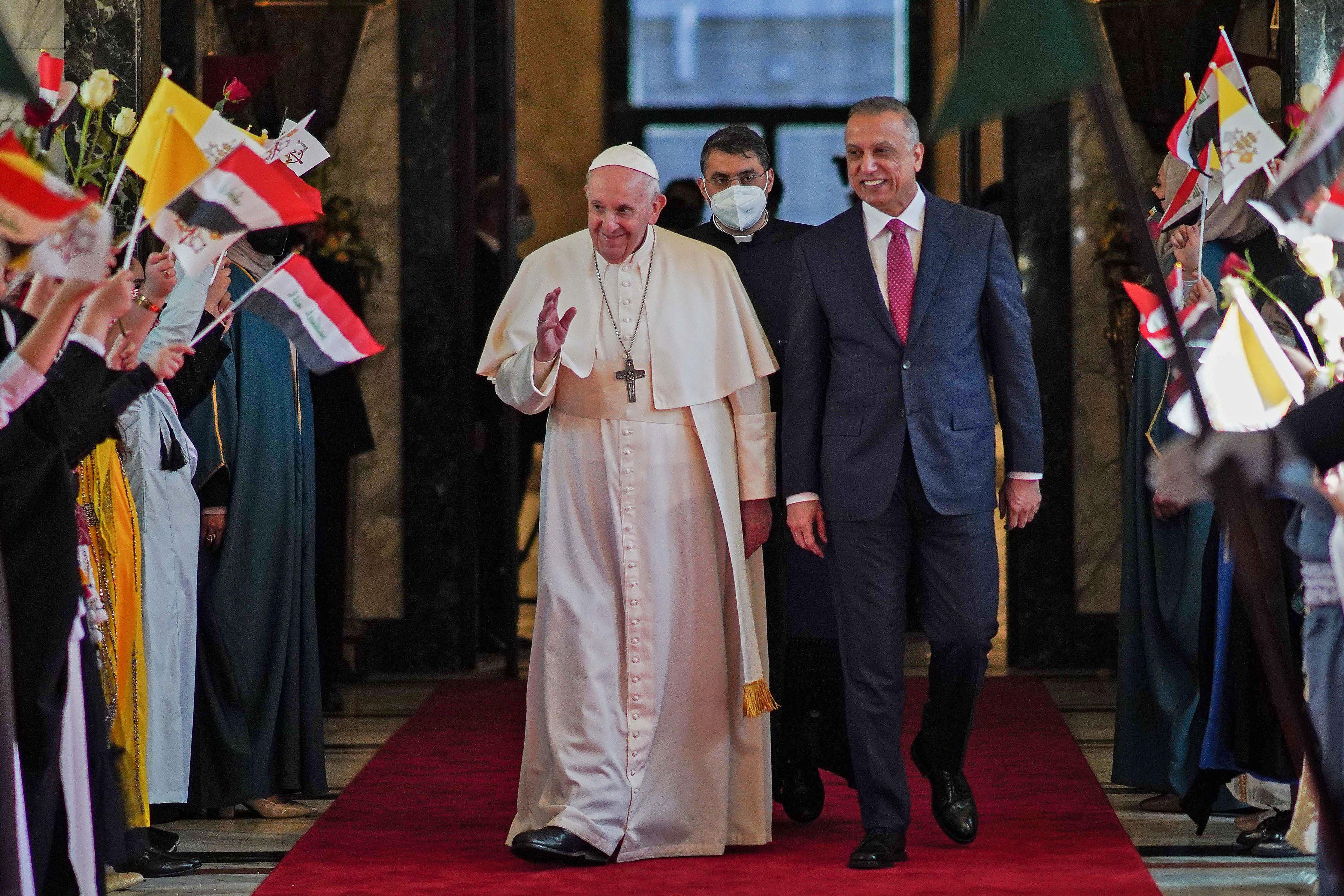 Pope Francis and Iraq's Prime minister walk down an aisle lined with people greeting them with flags.