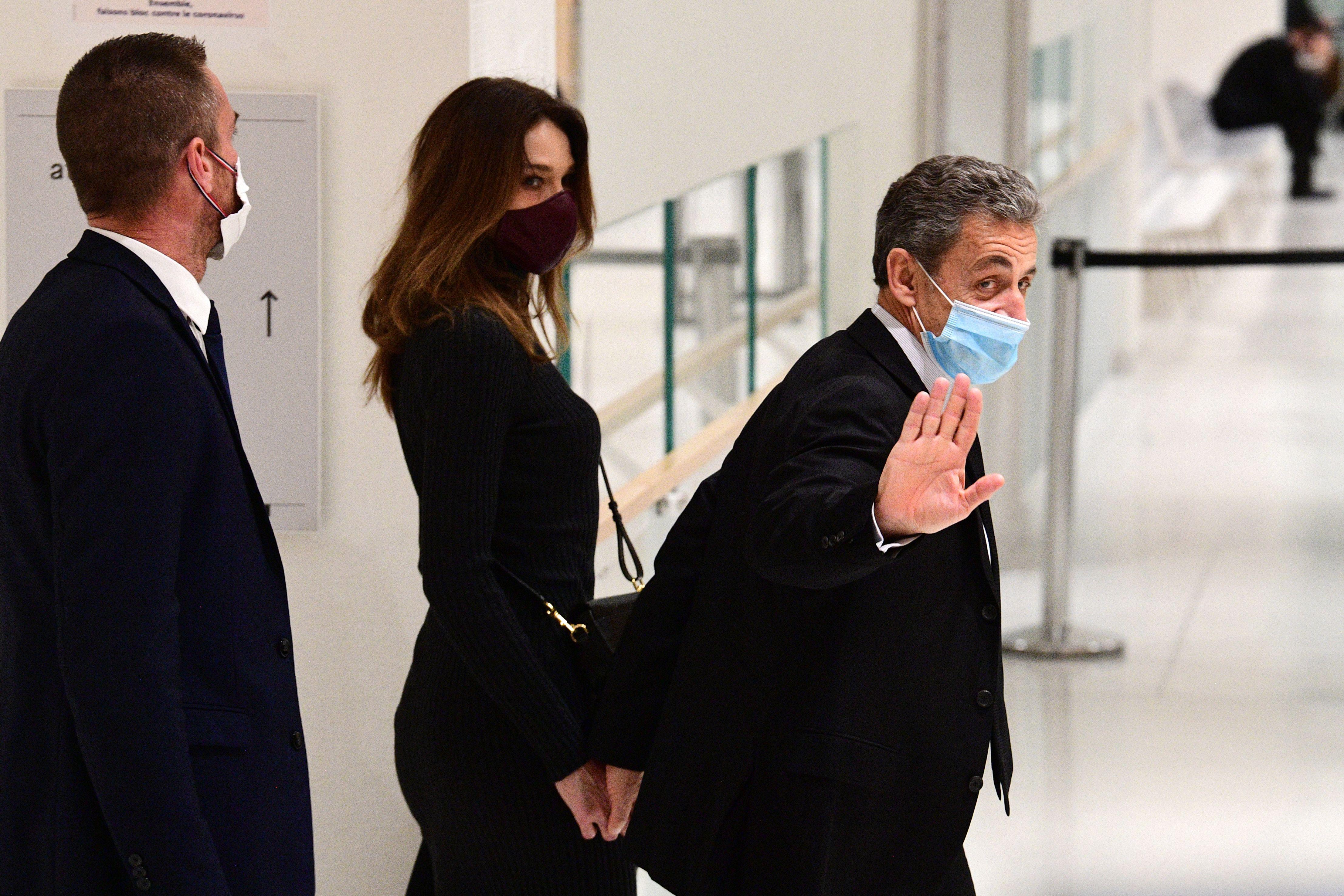 Sarkozy waves to the camera with one hand and holds his wife's hand with the other as they walk through a hallway
