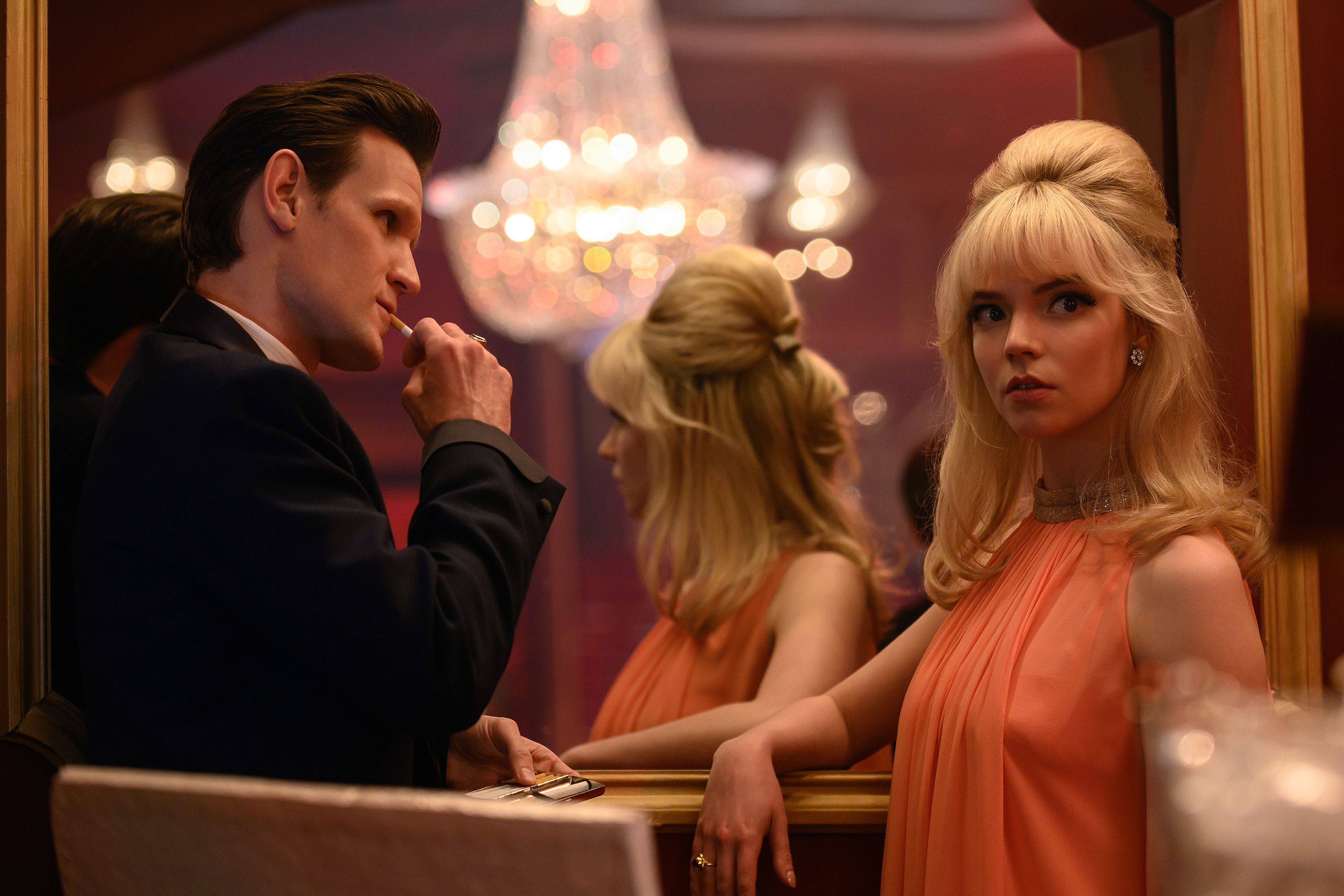 A dapper looking man in a suit jacket with his hair slicked back lights a cigarette while standing near and looking entranced with a blonde woman, dressed in 1960s attire and up-do hair style, looks away from him.