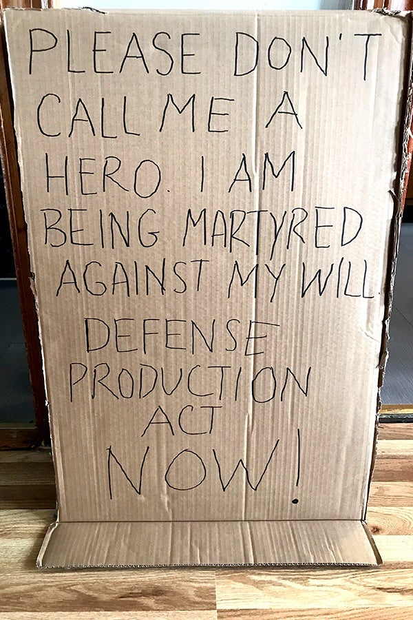 A cardboard sign that says "PLEASE DON'T CALL ME A HERO. I AM BEING MARTYRED AGAINST MY WILL. DEFENSE PRODUCTION ACT NOW!"