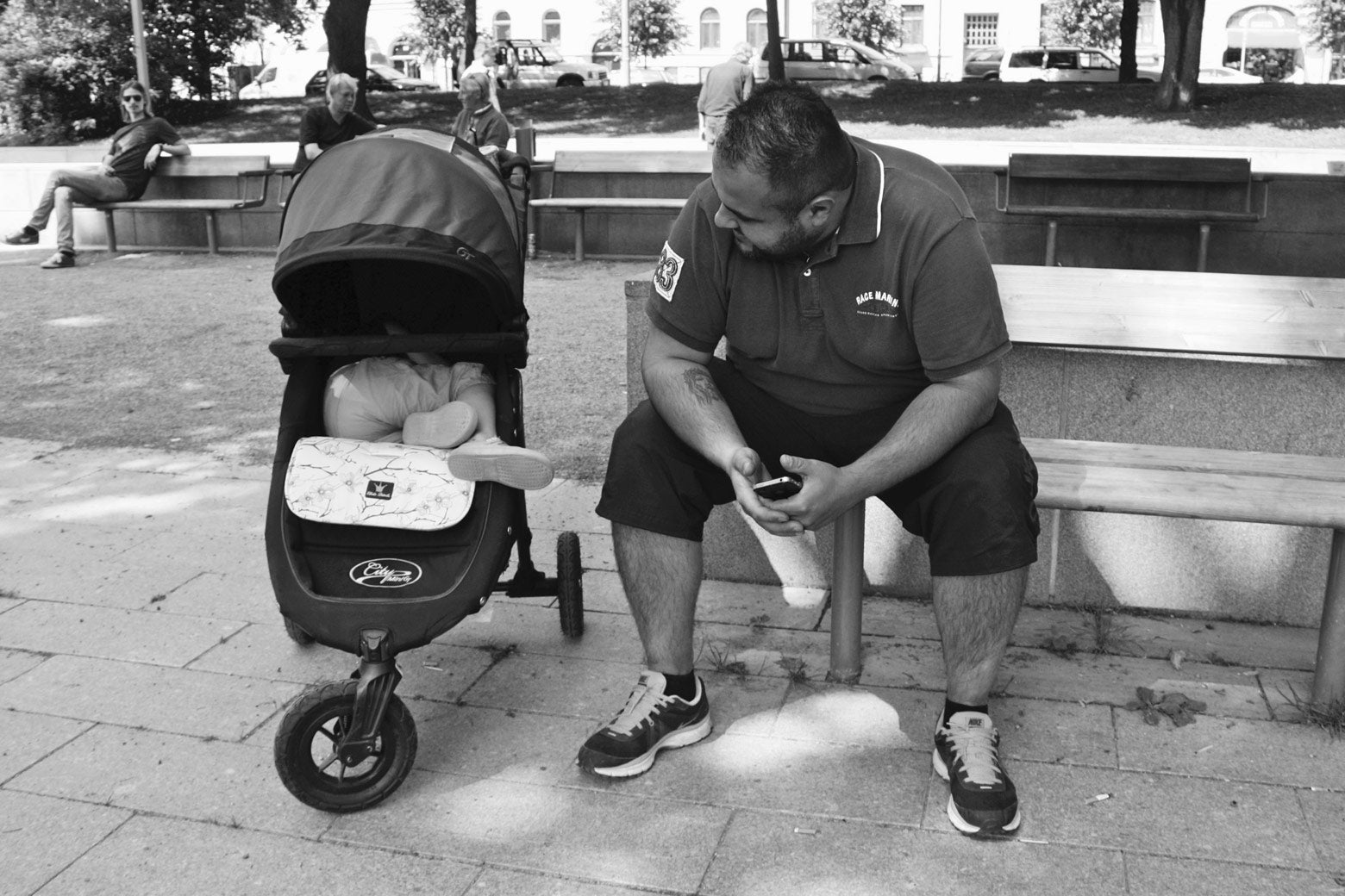 A father stops for a break on a park bench while his daughter naps in her stroller.