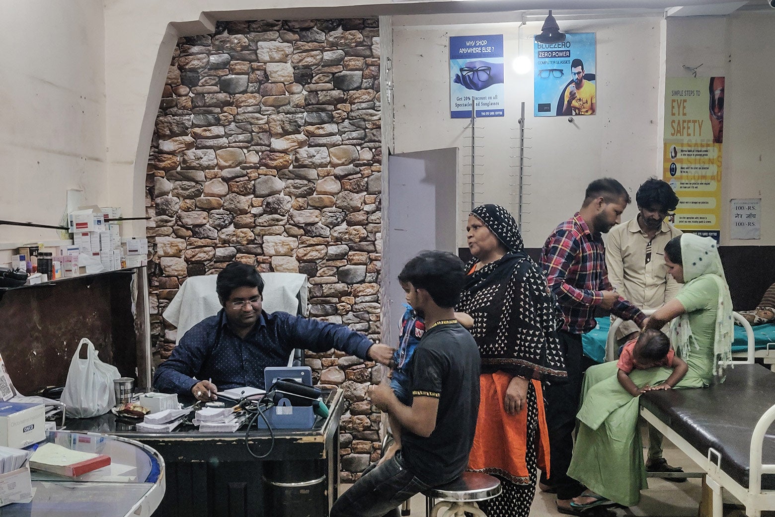 A doctor conducts an examination of a toddler in a cluttered consult room. Behind him, five other patients wait.
