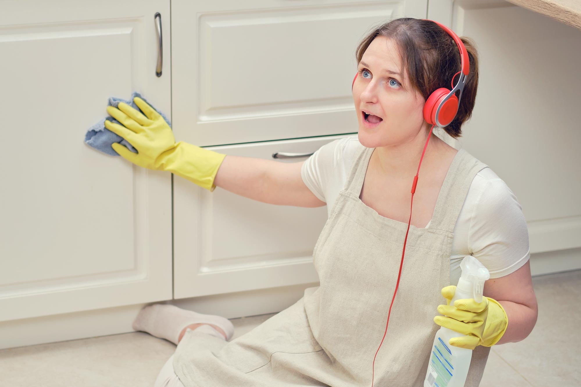 A woman wearing red headphones and yellow rubber cleaning gloves. She sits on the floor, wiping down a cabinet while looking enthralled by whatever she's listening to.