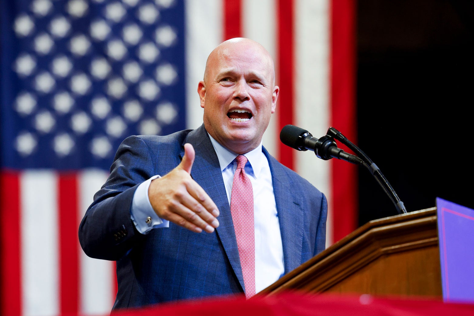 Whitaker speaking into a microphone at a podium, gesturing with his right hand outward, with an American flag hanging behind him