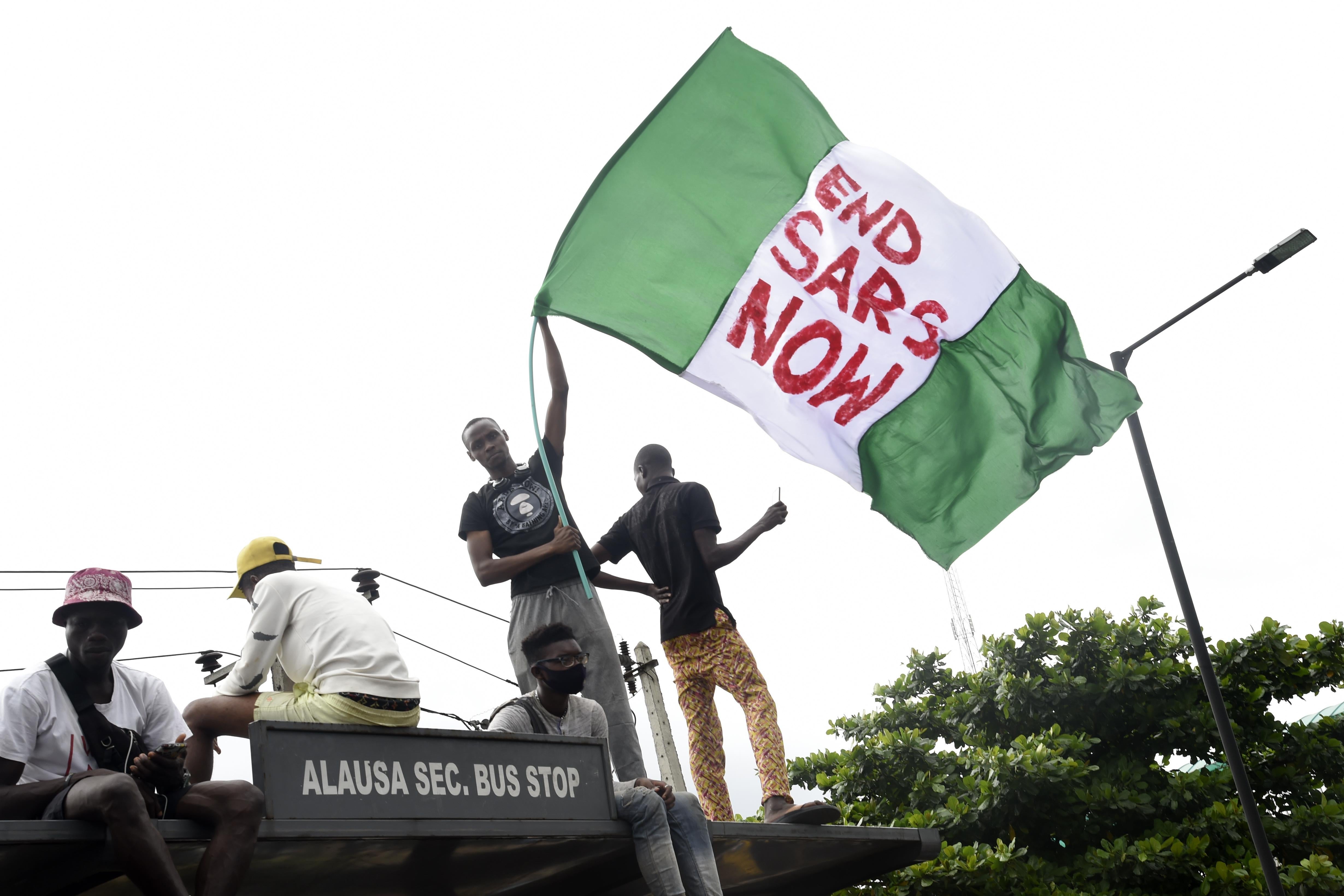 A protester standing on top of a bus stop with others waves a giant Nigerian flag that says "End SARS Now."
