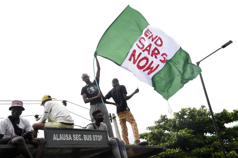 A protester standing on top of a bus stop with others waves a giant Nigerian flag that says "End SARS Now."