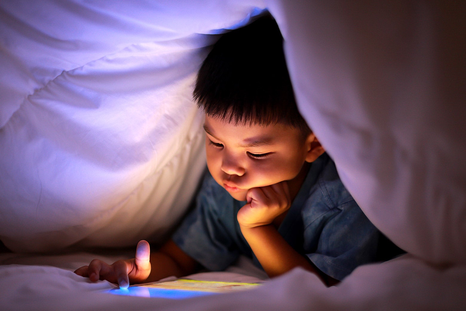 A small child uses a tablet under the covers.