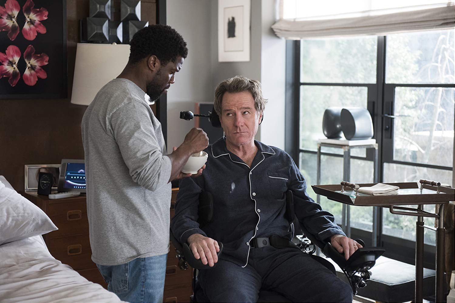 Kevin Hart holds a bowl and stands in front of Bryan Cranston, seated in a wheelchair.