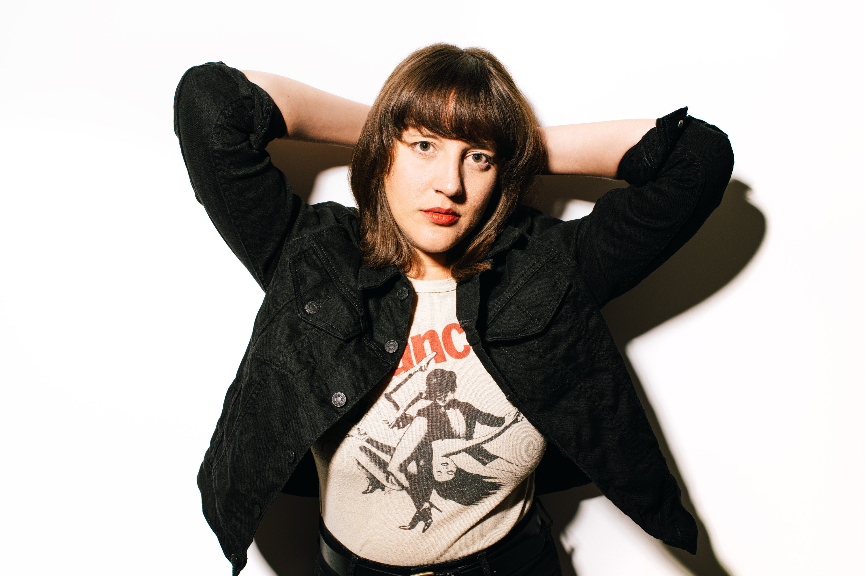 A woman with short brown hair stands against a white wall, wearing a Bob Fosse T-shirt.