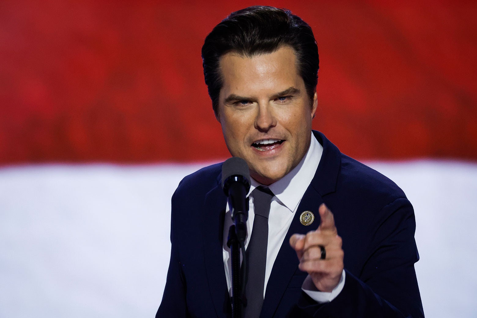 Gaetz speaks into the microphone while looking and pointing into the camera, an American flag behind him