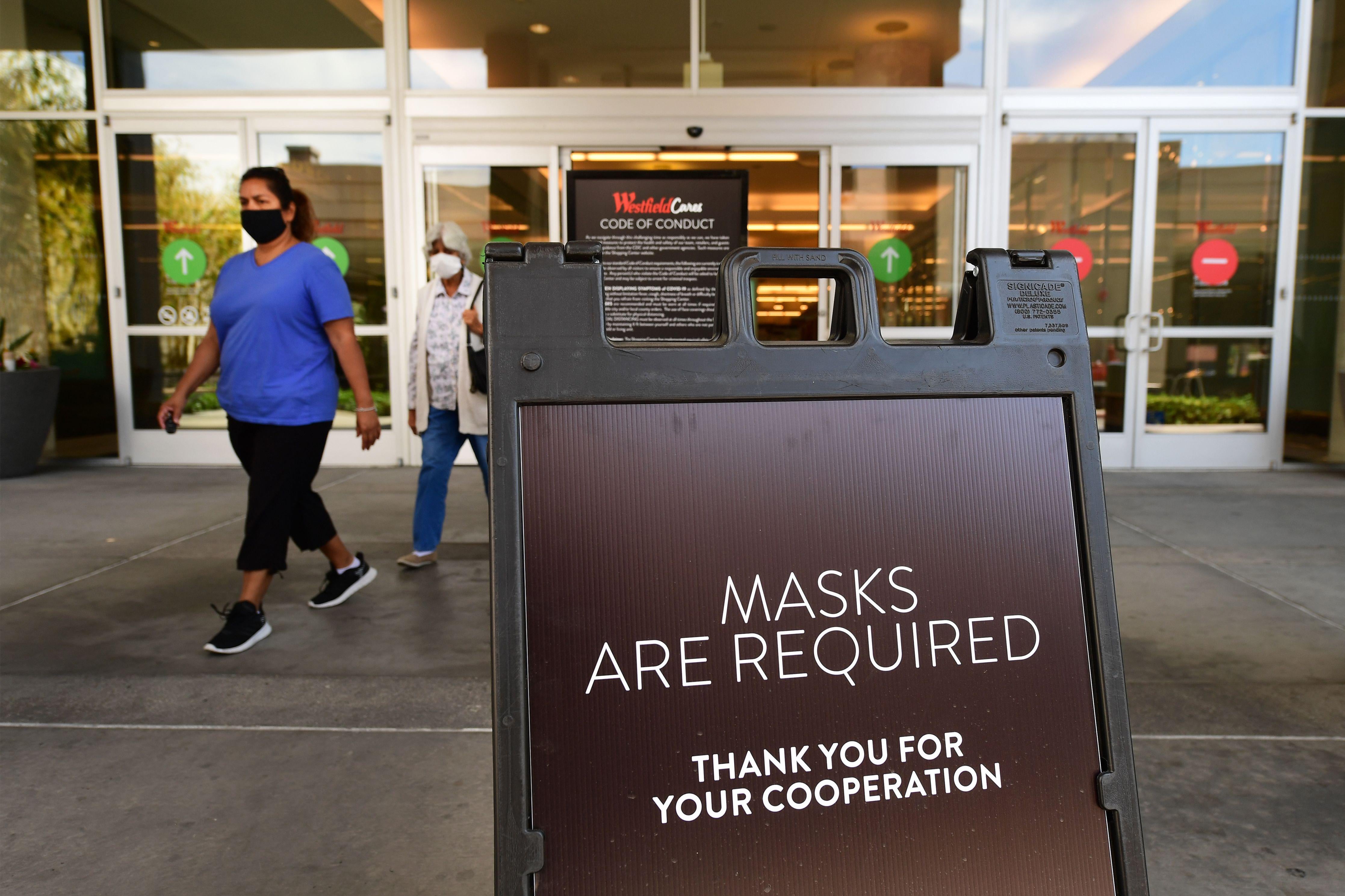 Women wearing face masks walk out of mall doors, by a sign that says "Masks Are Required."
