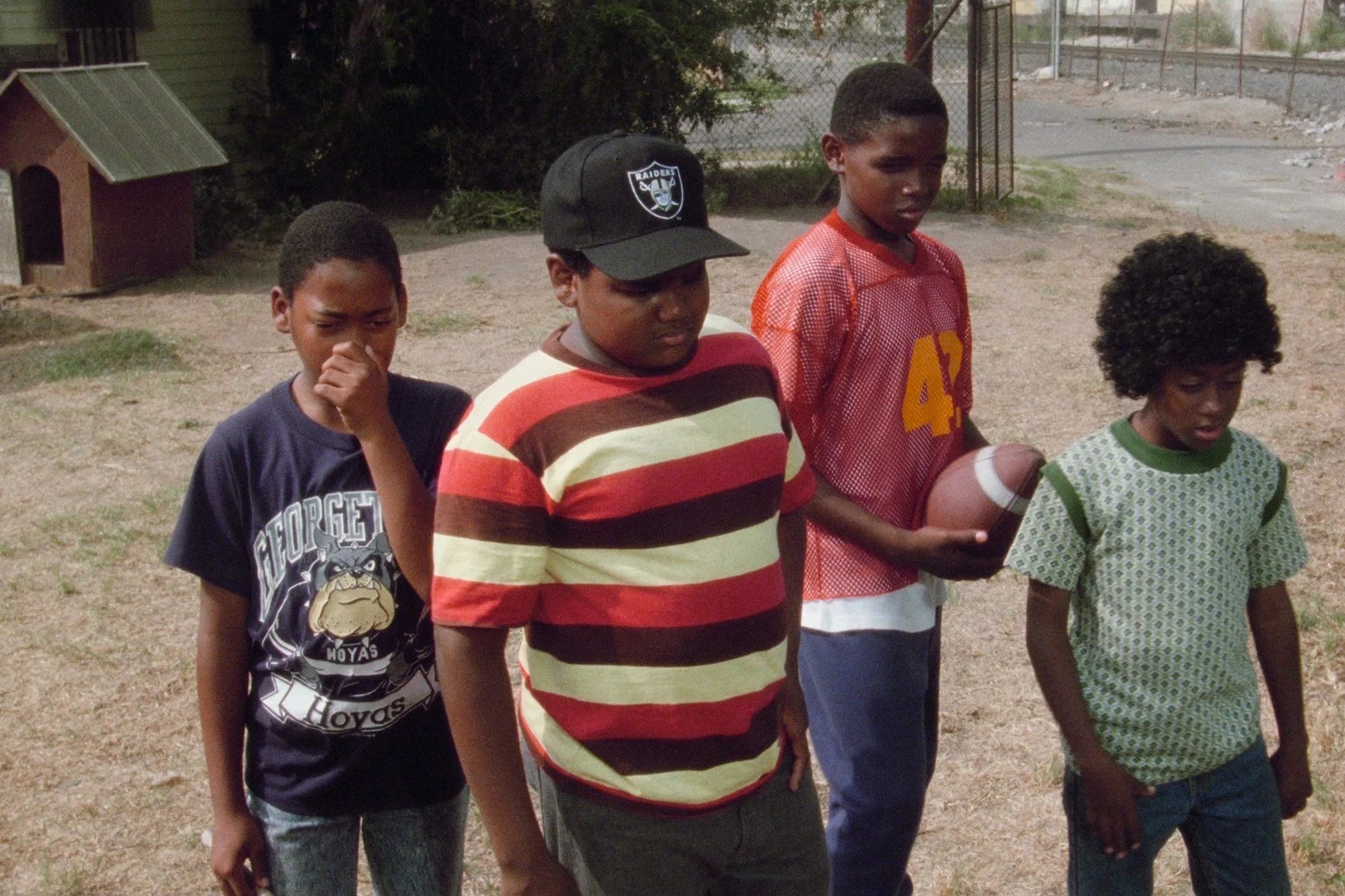 Four young boys stand while looking down at something off camera with frowns or straight faces. From left to right: Desi Arnez Hines II holds his nose, Baha Jackson sports a frown, Donovan McCrary looks down while holding a football, and Kenneth A. Brown looks down.