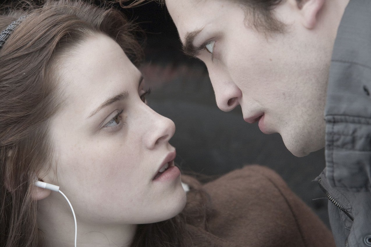 A girl with brown hair and wearing white earbuds looks up at a man with brown hair, who stands very close to her face. 