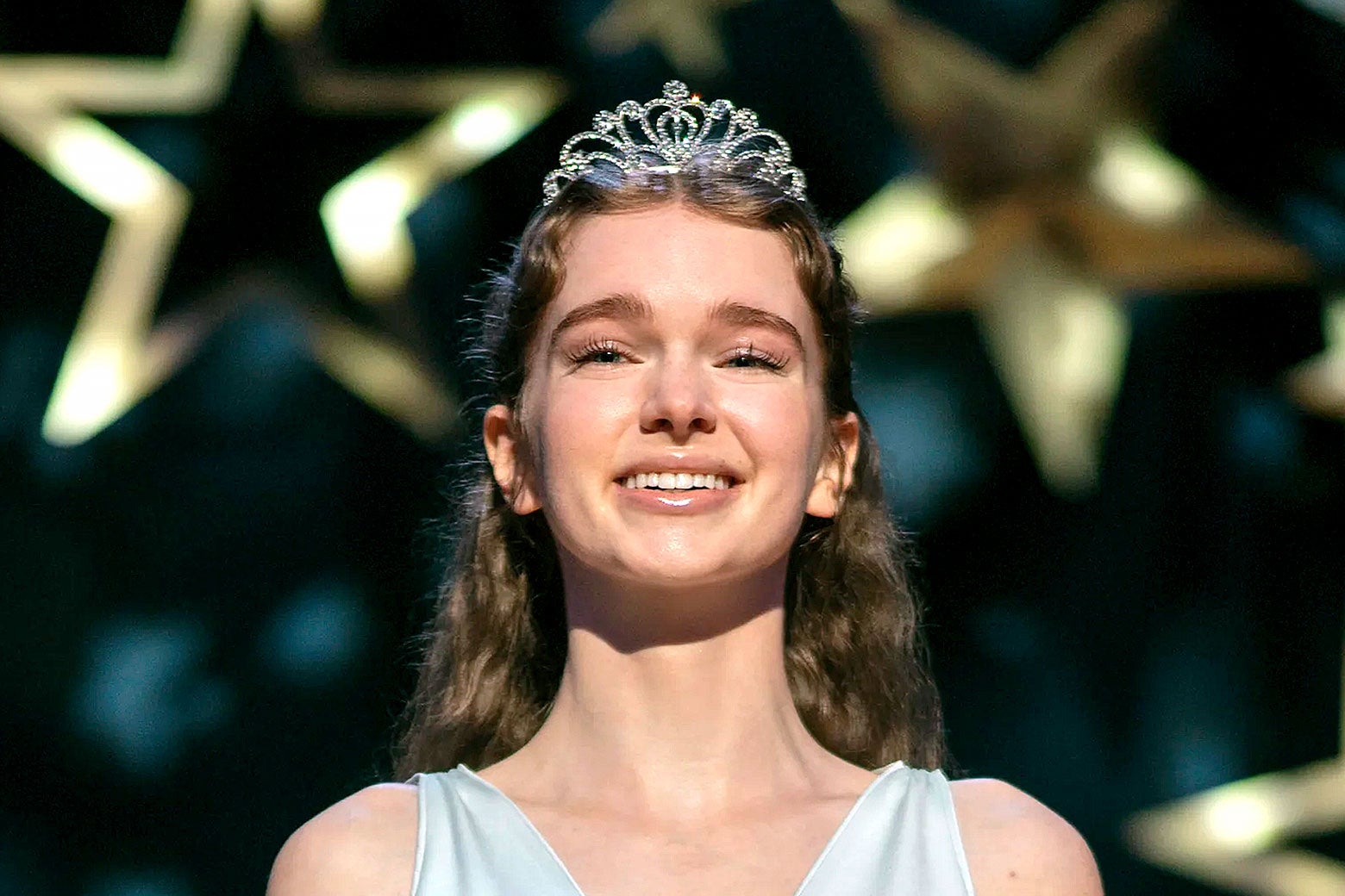 A girl smiling on stage in a dress and a prom queen crown.