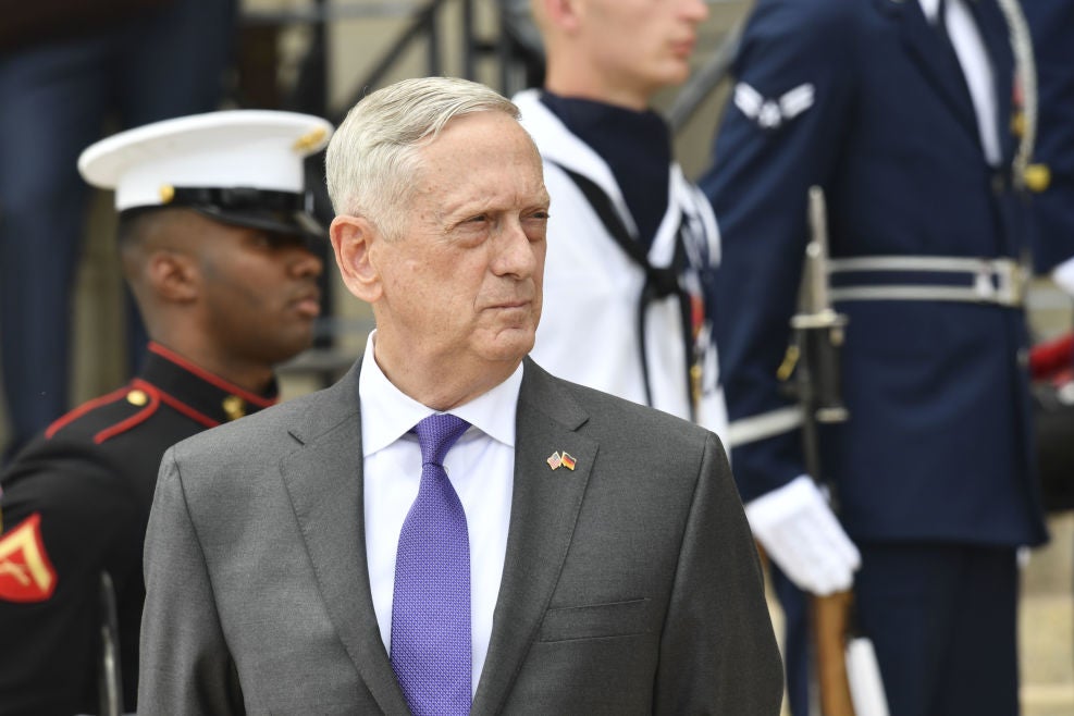 Mattis, wearing a suit and backdropped by several in-uniform service members, looks to his left.