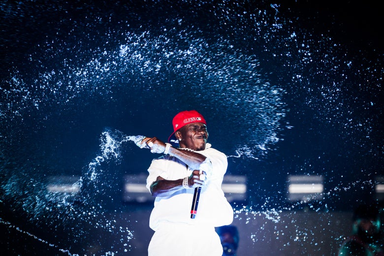 A man dressed in all white and wearing a red hat throws water across a stage as he holds a microphone. 