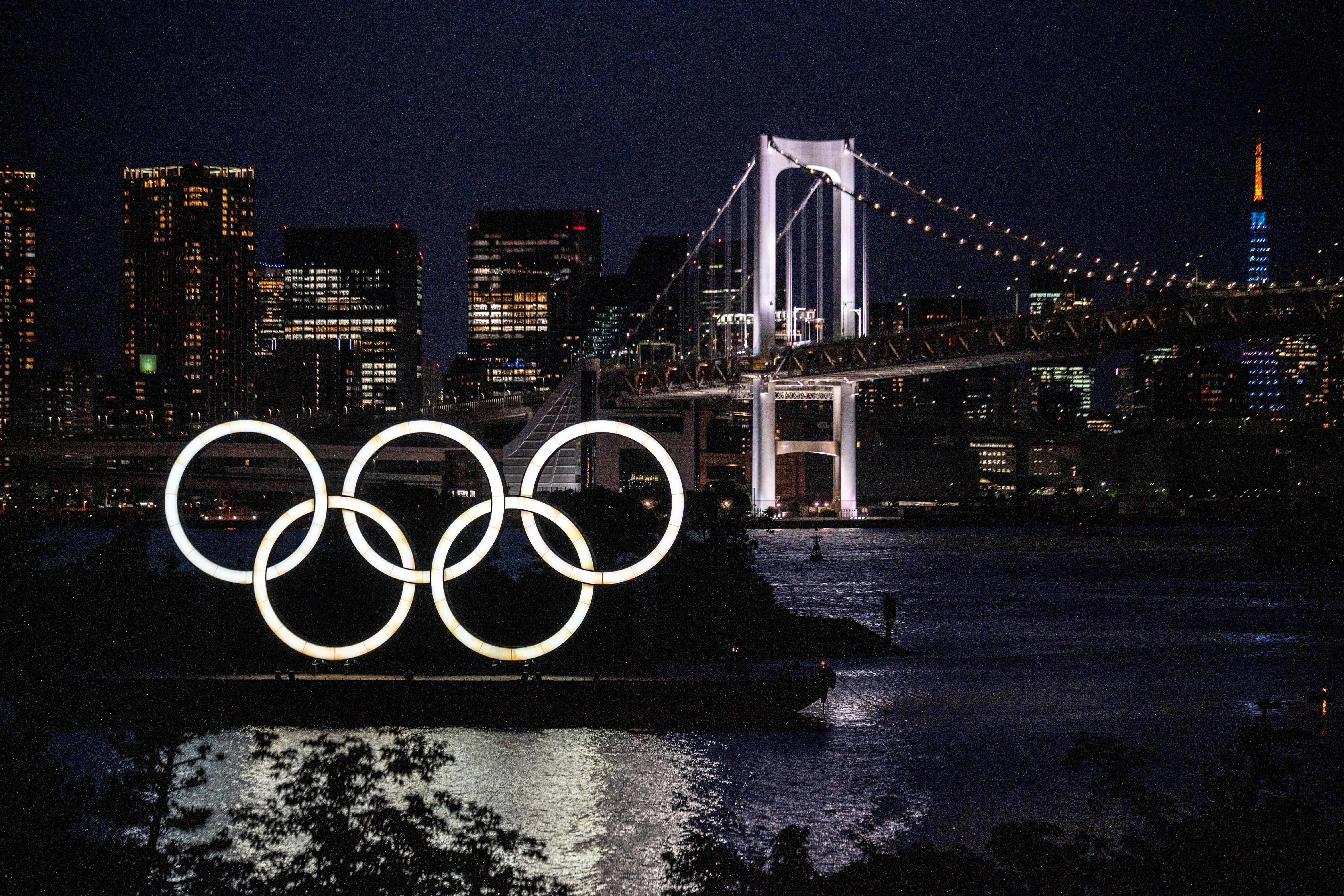 The Olympic rings are seen lit up at a waterfront, in front of a bridge and the Tokyo skyline.