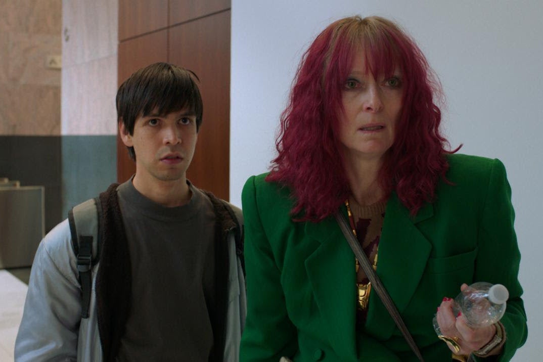 Torres looks boyish in a brown moptop, a hoodie, and a backpack. Swinton meanwhile wears a bright green blazer and pink hair that looks like a too-old dye job. They stand looking concerned toward the camera in a city lobby.