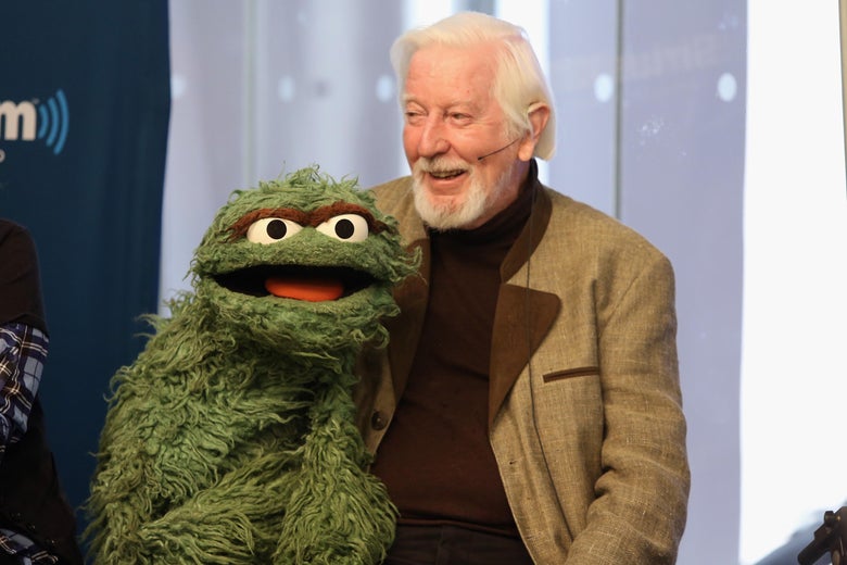 Caroll Spinney, wearing a light brown suit jacket, holds the Oscar the Grouch puppet.
