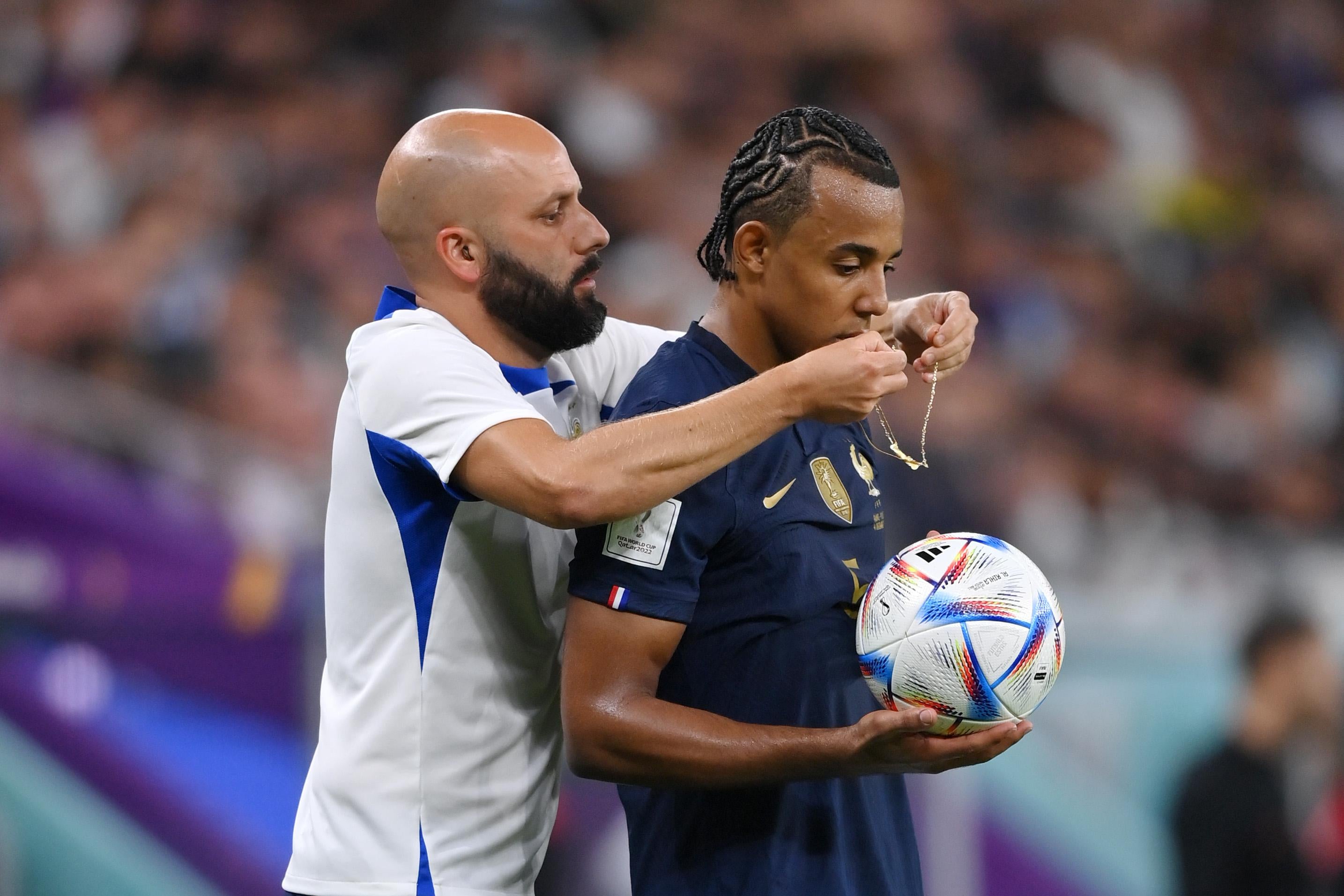 Jules Kounde holds the ball while an official stands behind him and removes a necklace from Kounde's neck.