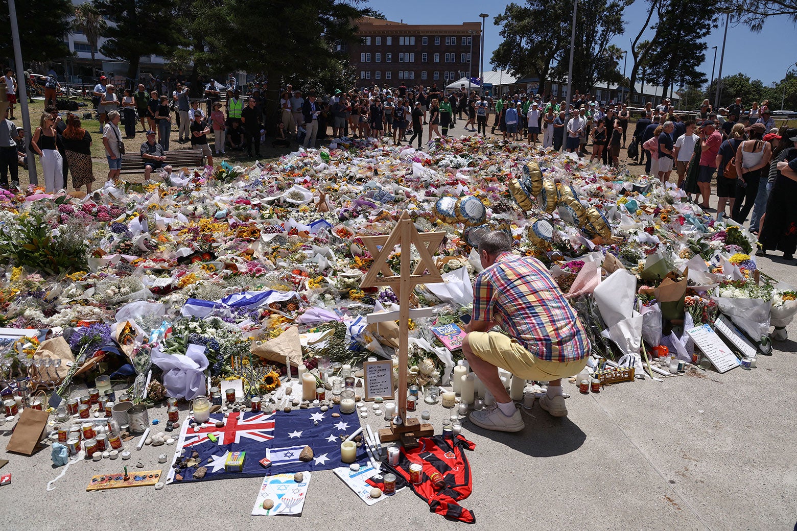 Piles of gifts, flowers, and signs on the beach surrounded by visitors; one man is lighting a memory candle in the foreground.