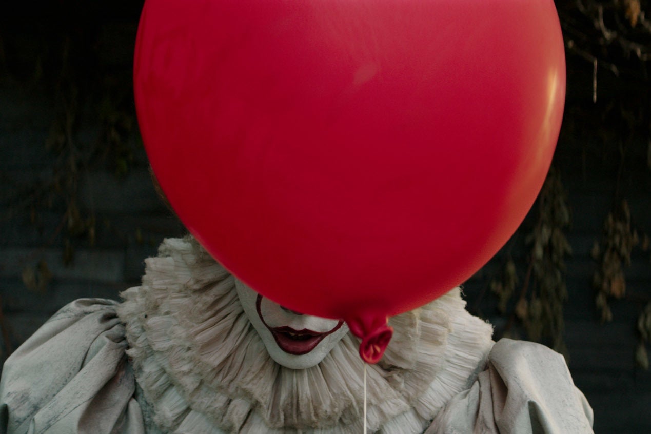 A clown dressed in white with red lip make-up hides most of his face above his mouth with a big red balloon.