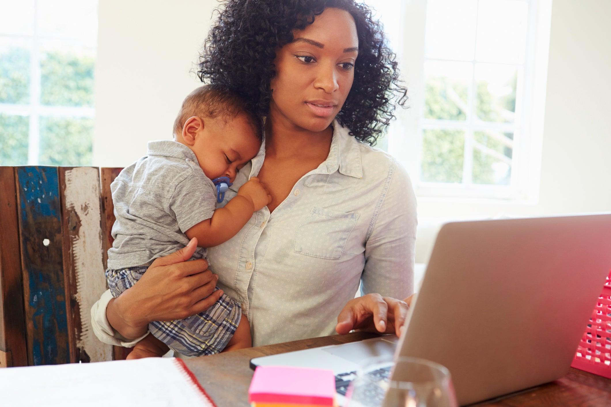 A woman holds a baby while working at her laptop.