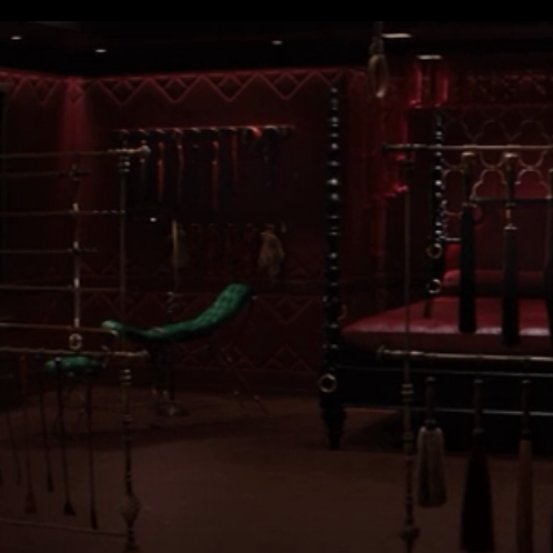 A red room with black accents, with a bed at the center and various floggers hanging