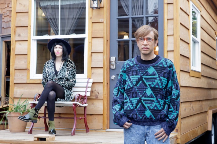 Carrie Brownstein (left) and Fred Armisen (right) stand morosely in character for Portlandia. Armisen wears a tragic, baggy hipster sweater and Brownstein is decked out in a wide-brim hat and cheetah-print wrap dress.