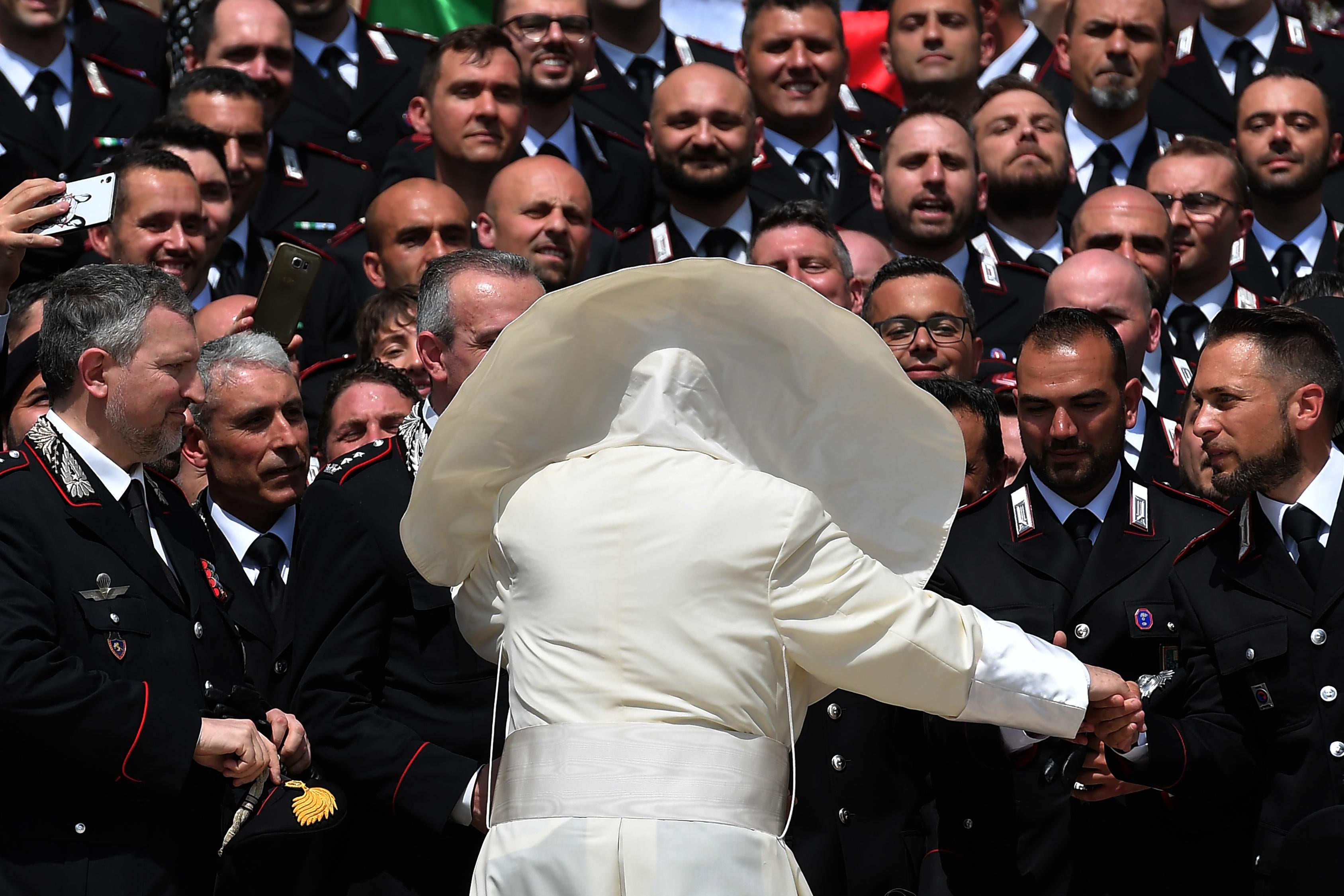 Pope Francis, seen from behind with his cape billowing in the wind, greets a large group of uniformed Carabinieri.