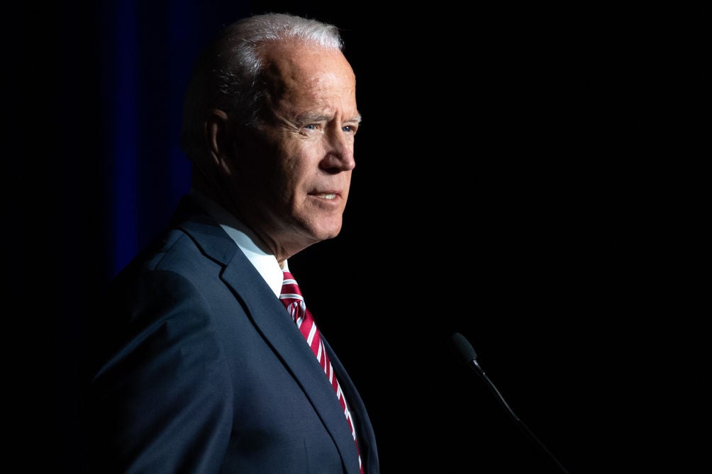 Biden, seen from the side, speaks into a microphone against a dark background.