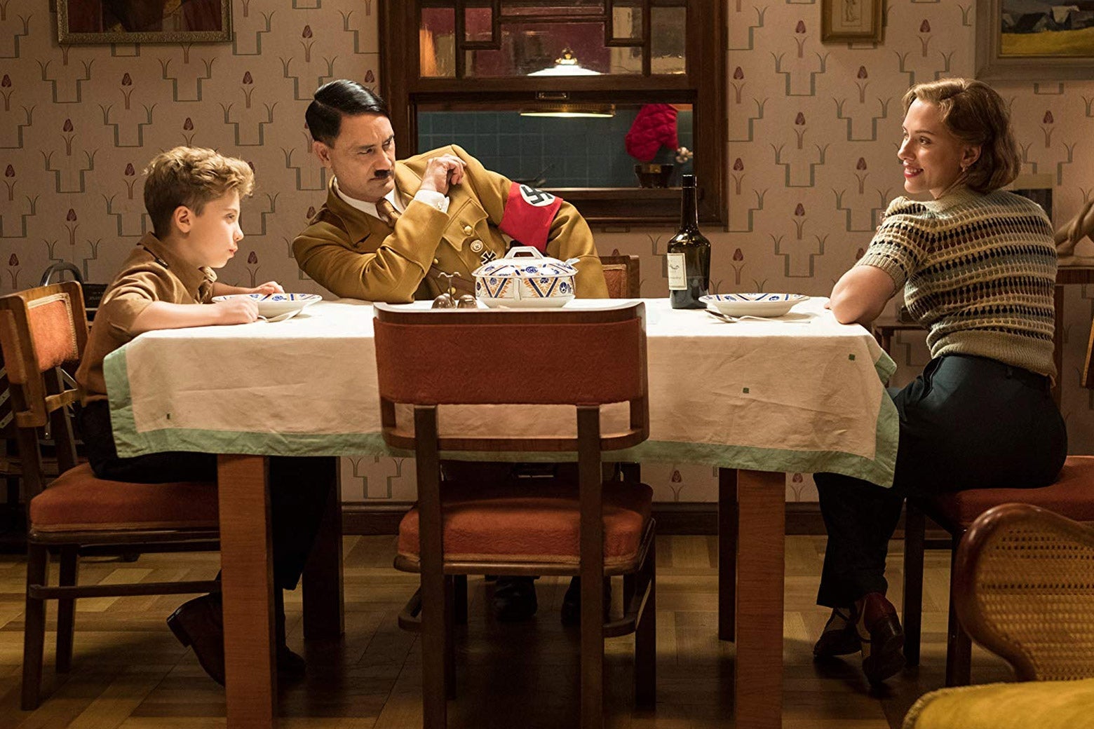 Roman Griffin Davis, Taika Waititi (as Hitler), and Scarlett Johansson sit around a table in a still image from the film.