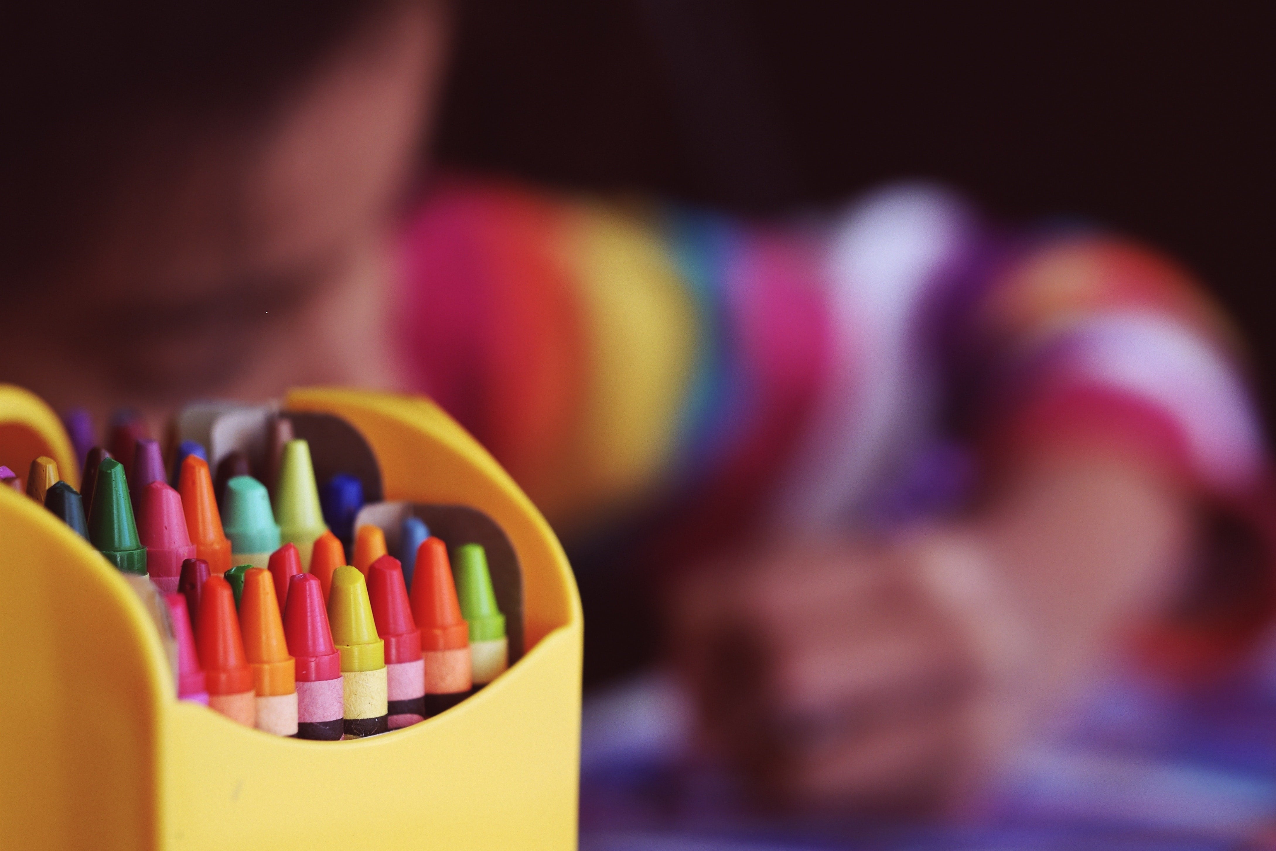 A box of crayons in focus, with a blurred child drawing in the background