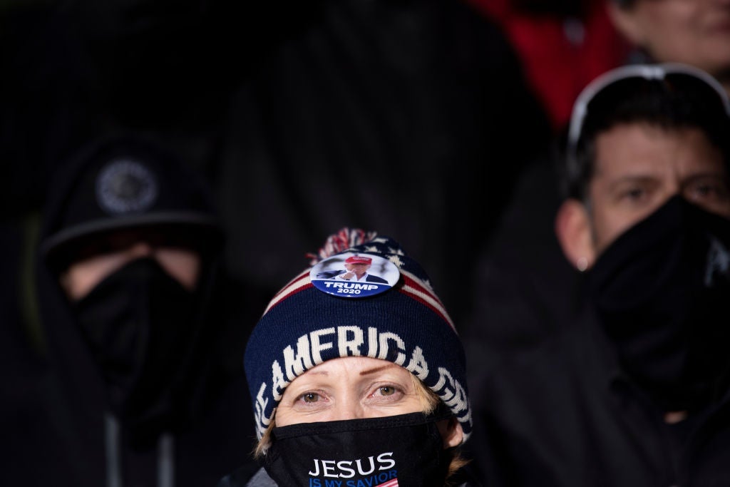 A woman wearing a Keep America Great beanie and a mask with "Jesus" written on it looks into the camera. Two men wearing masks and dark clothes stand behind her.