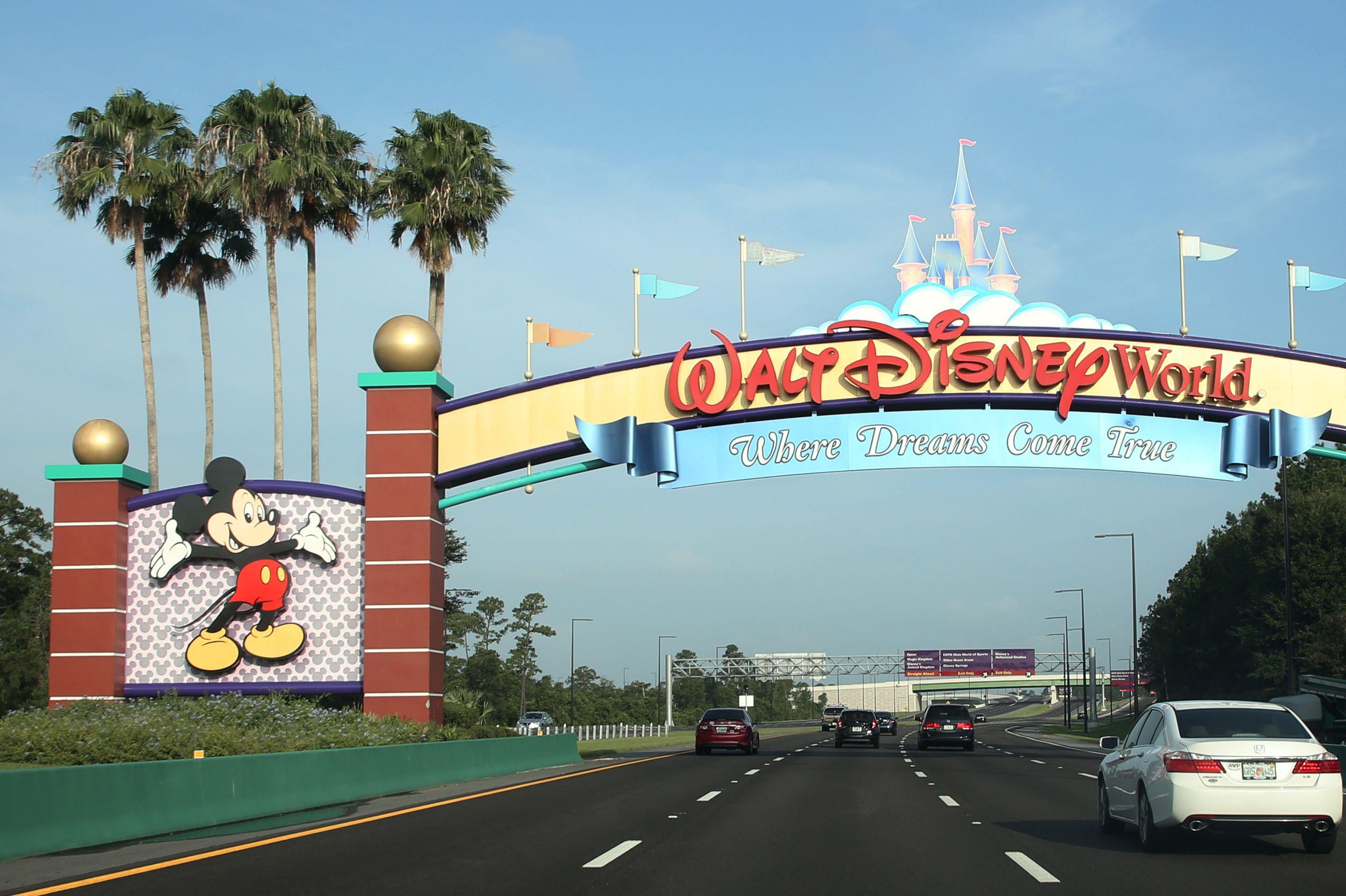 A sign that says "Walt Disney World: Where Dreams Come True" above a four-lane highway with palm trees and a clear blue sky in the background