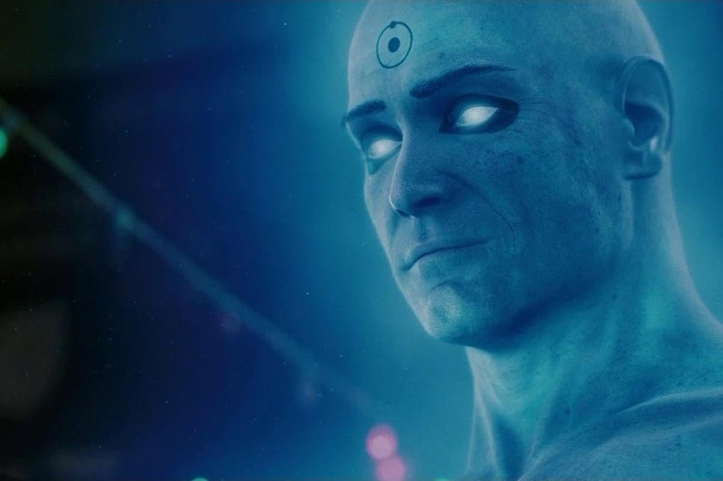 Billy Crudup in entirely blue make-up with Dr. Manhattan's signature symbol on his forehead stares off camera with glowing blue eyes. 