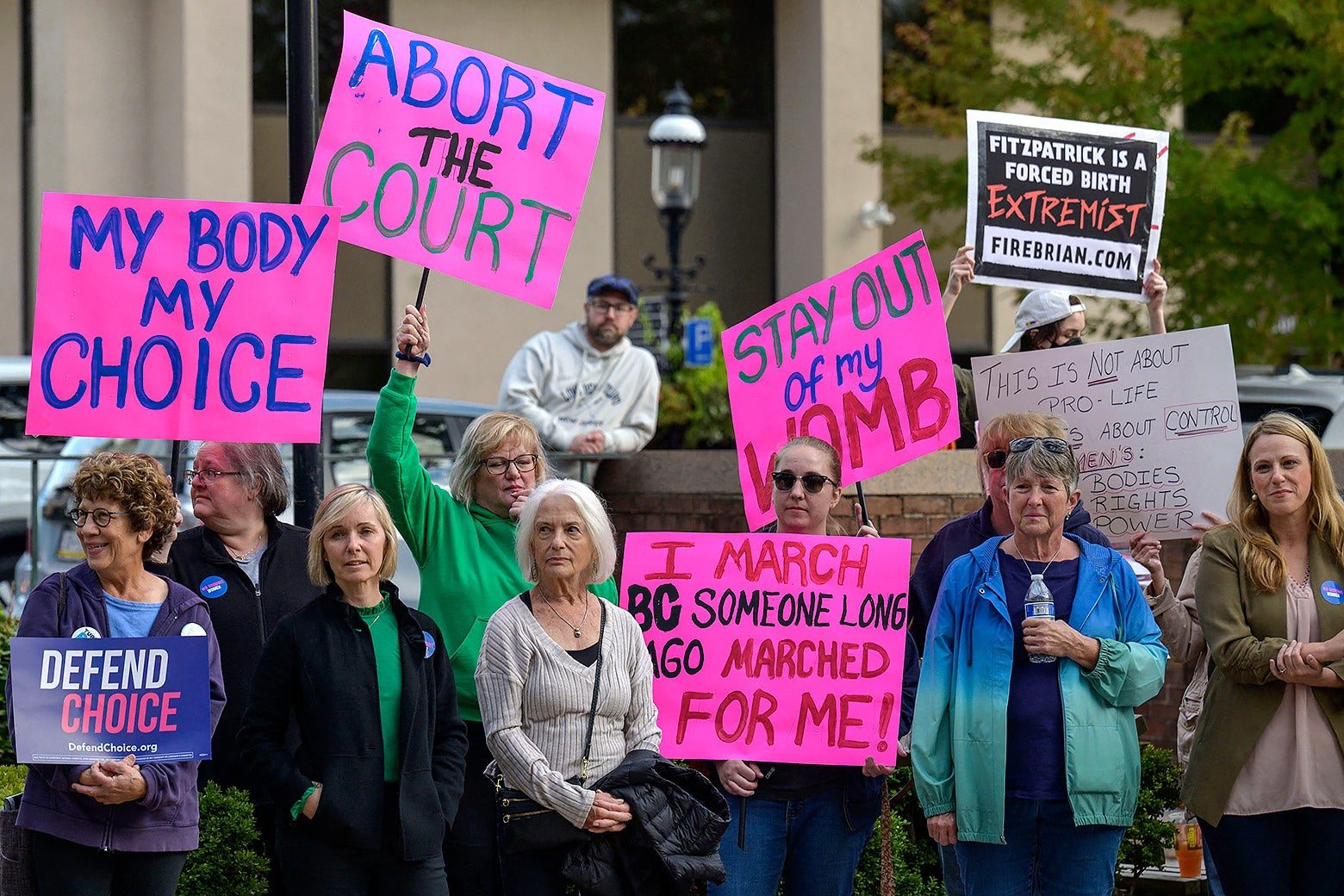 Protesters hold up signs that read "abort the court," "my body my choice," "stay out of my womb," and "I march bc someone long ago marched for me."