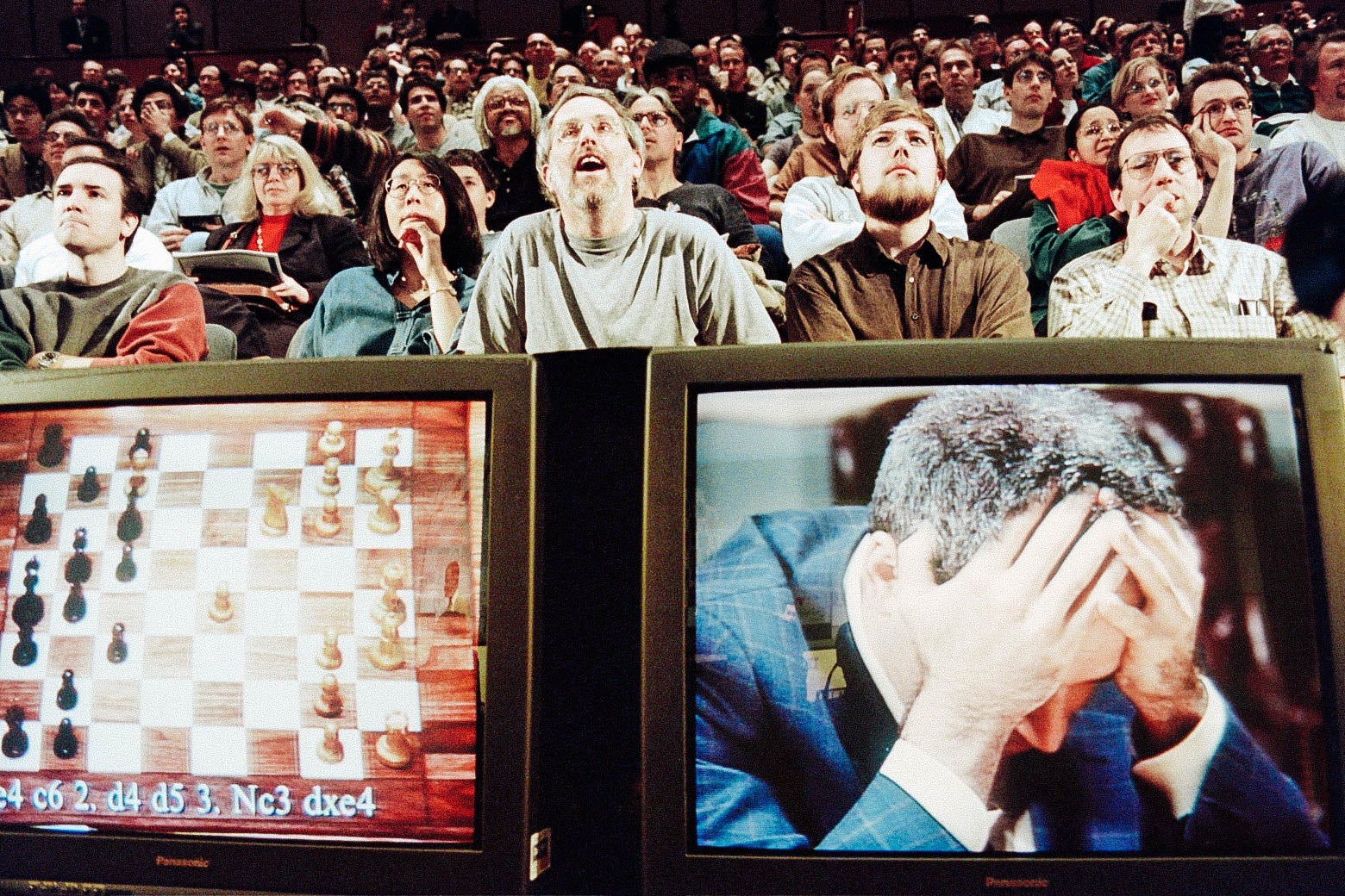 Two TV screens are seen below a sitting audience. The left screen shows a chessboard; the right shows Kasparov holding his head in his hands.