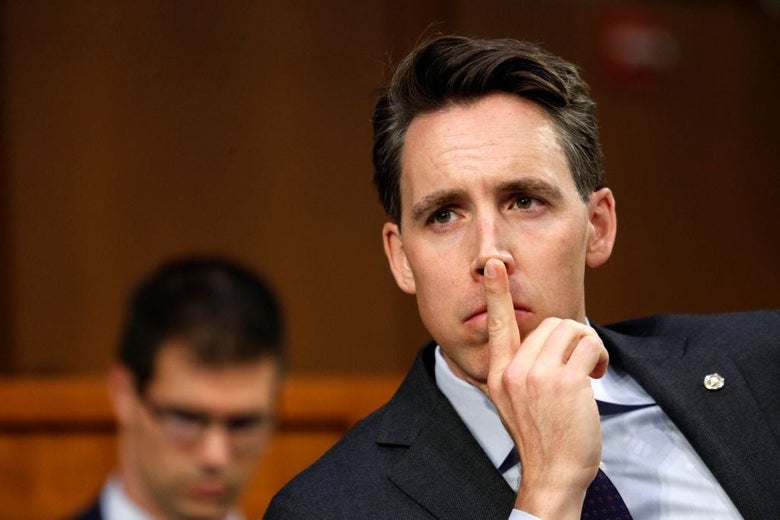 Josh Hawley, seated in a hearing room, presses his finger to the front of his nose.