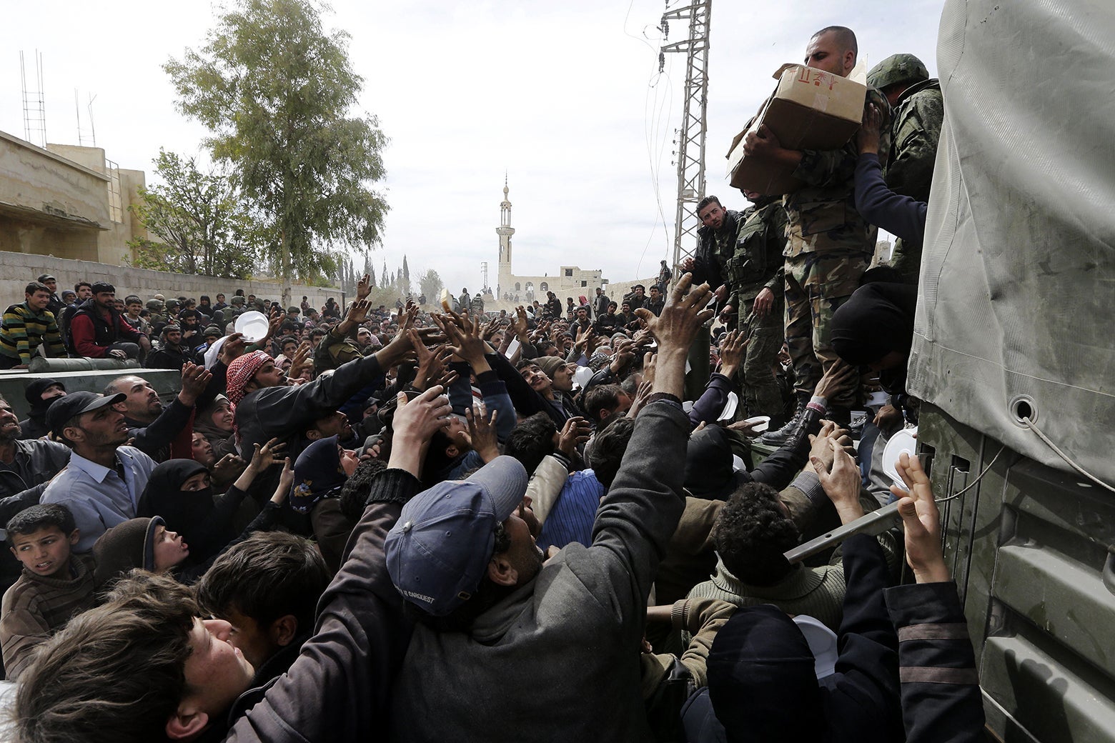 A crowd of Syrians stretching their arms up to soldiers holding boxes of food.
