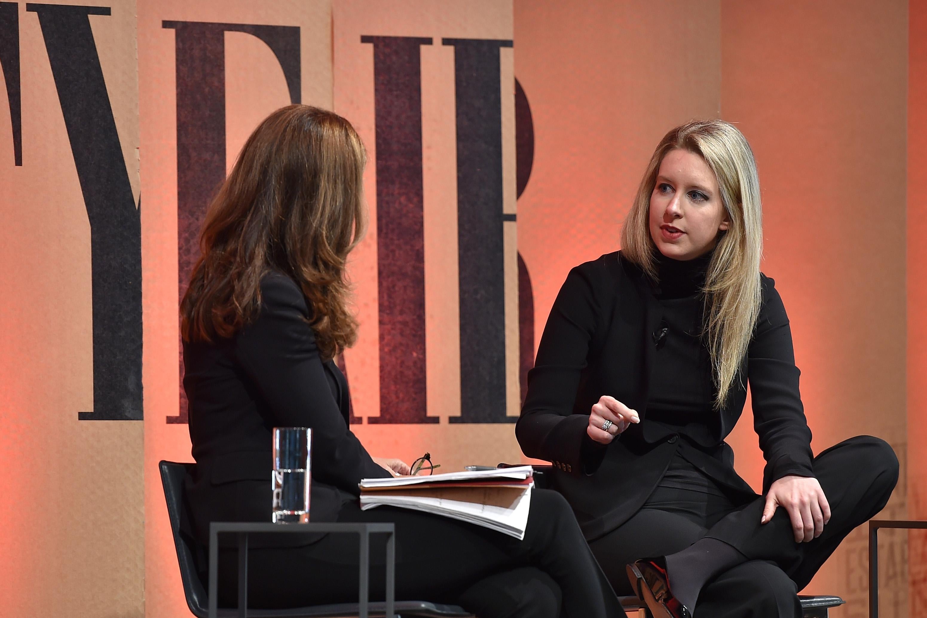 Elizabeth Holmes seated on a stage before an orange backdrop with Vanity Fair lettering