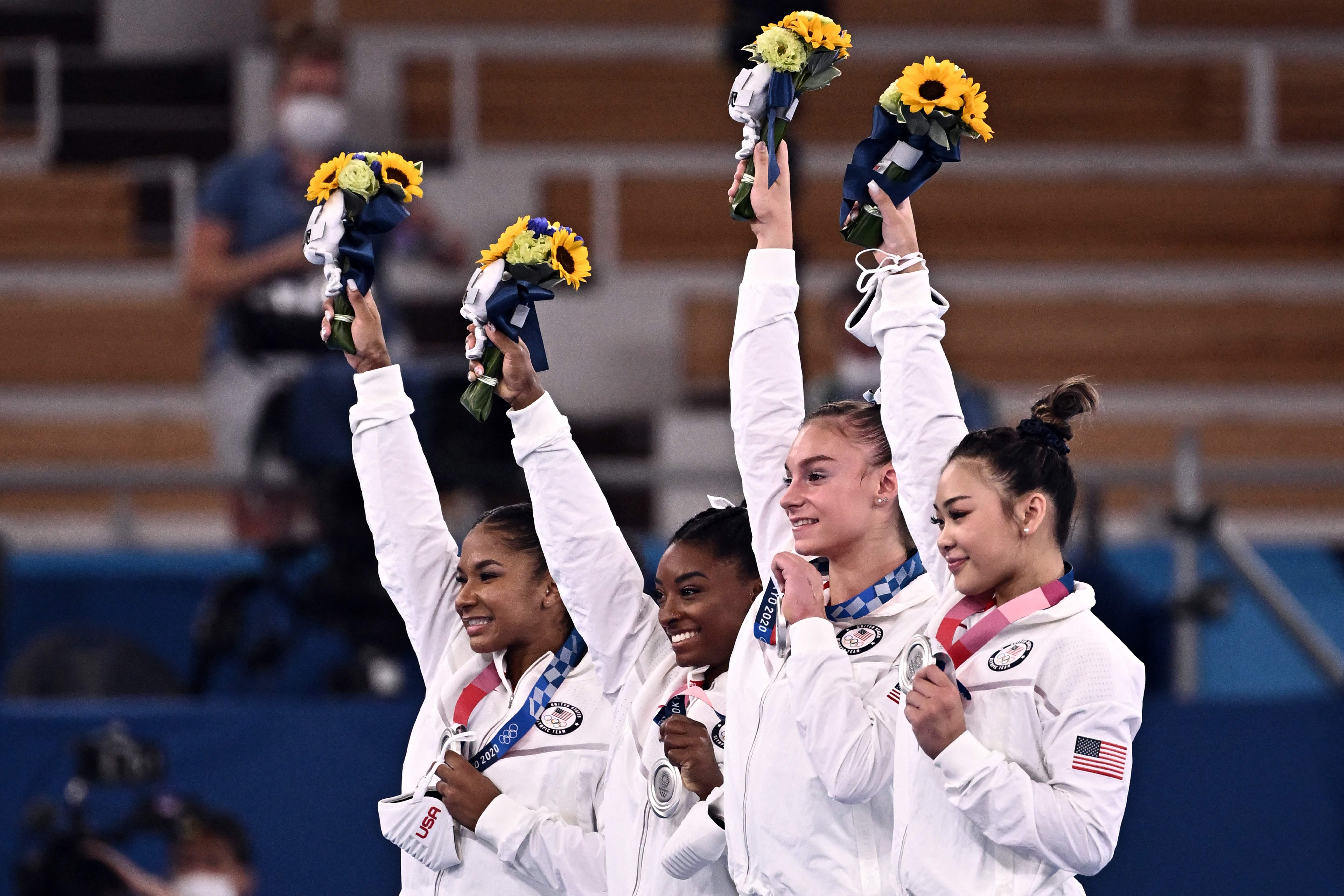 The four gymnasts stand in a row smiling on the podium. Each holds her bouquet up high with one hand and her silver medal around her neck with the other hand.