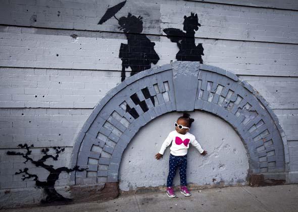 A child poses for a photo under a new art piece by British graffiti artist Banksy in the Brooklyn borough of New York, October 17, 2013.