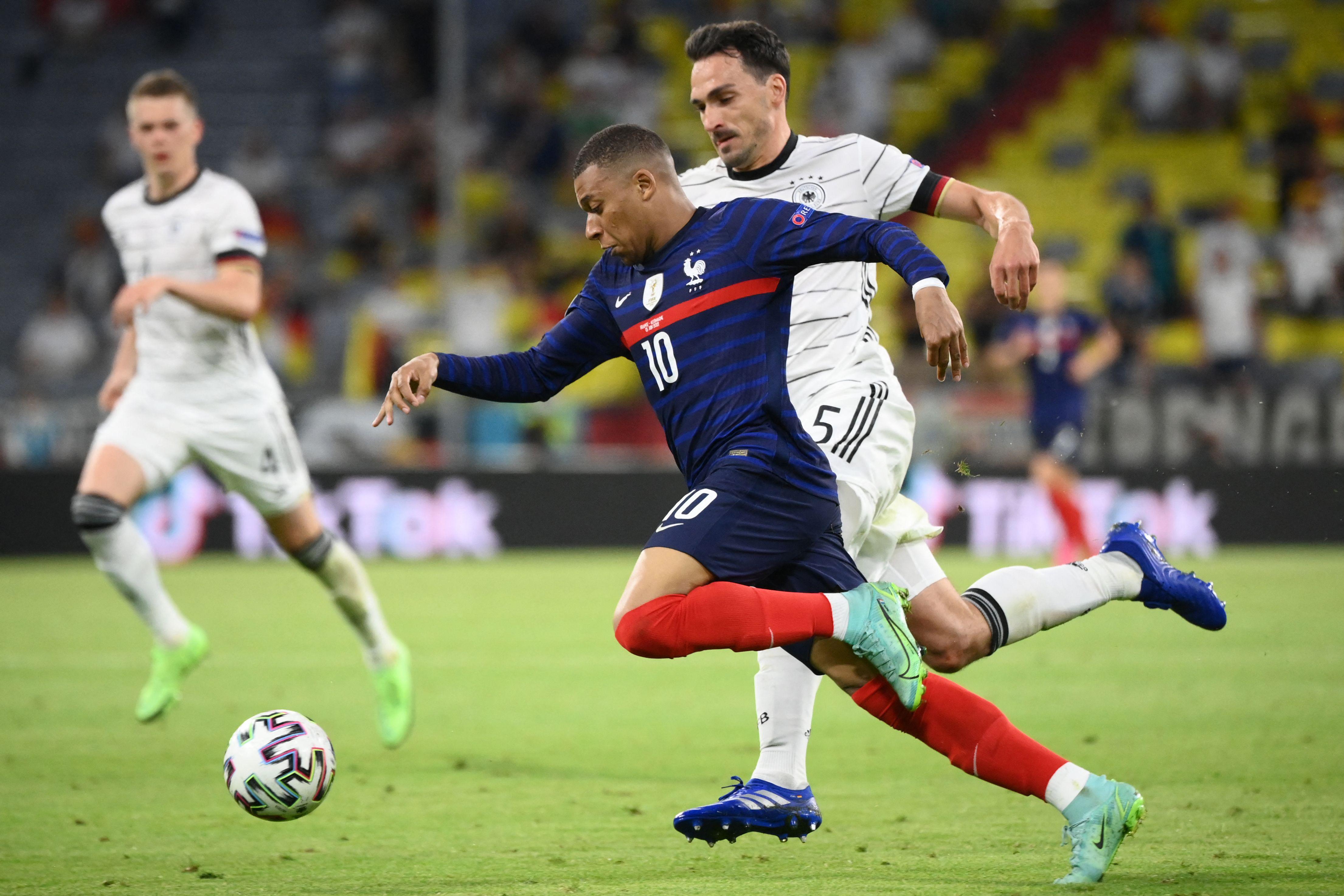 Kylian Mbappe chases the ball on the pitch against Germany, Mats Hummels chasing next to him, with another German player watching in the background