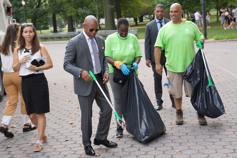 Eric Adams, in sunglasses and a suit, picks up trash while surrounding parkgoers carry large bags.