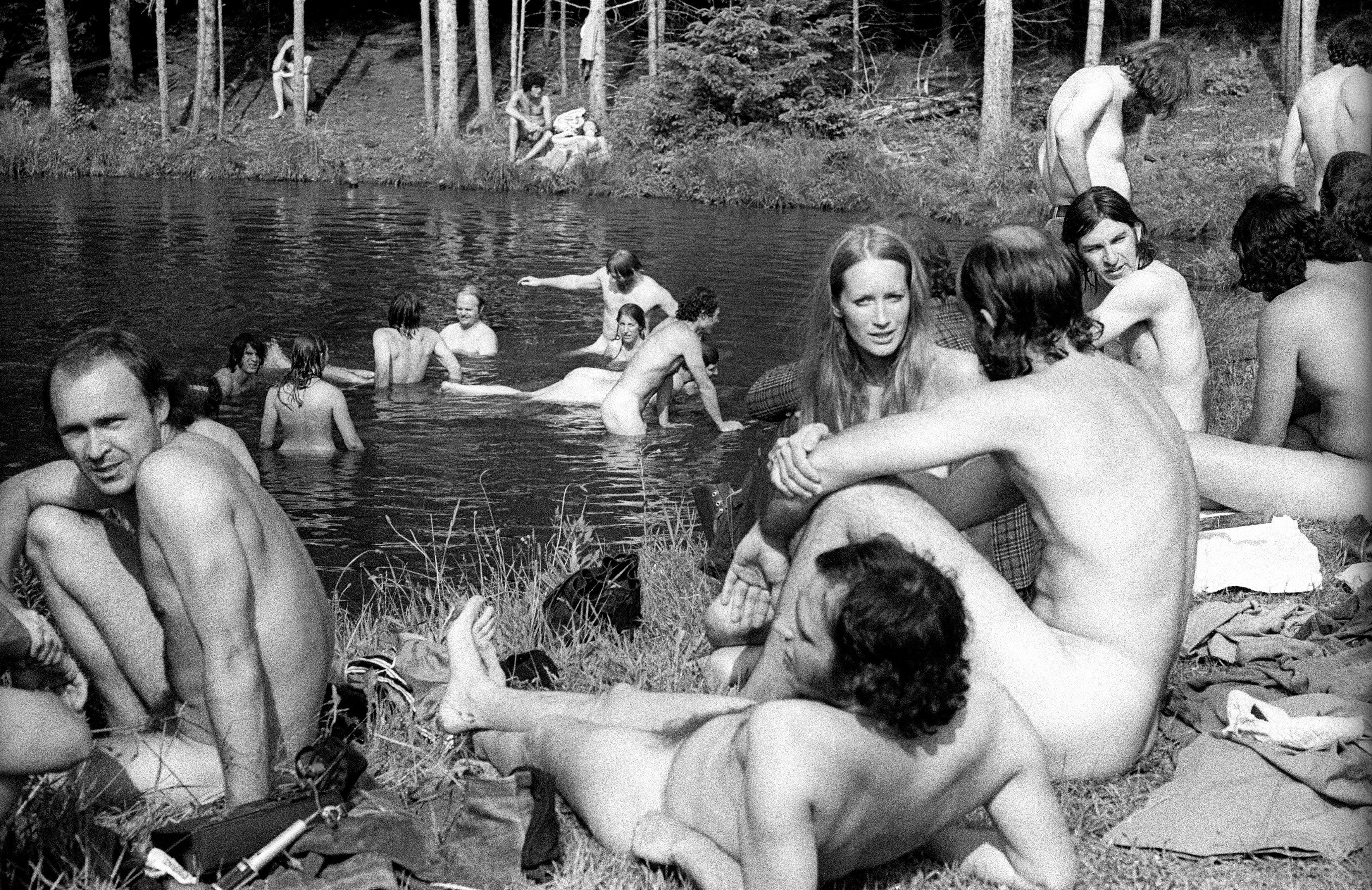 A 1970 black and white photo of nudists bathing in a river and sitting on the banks.