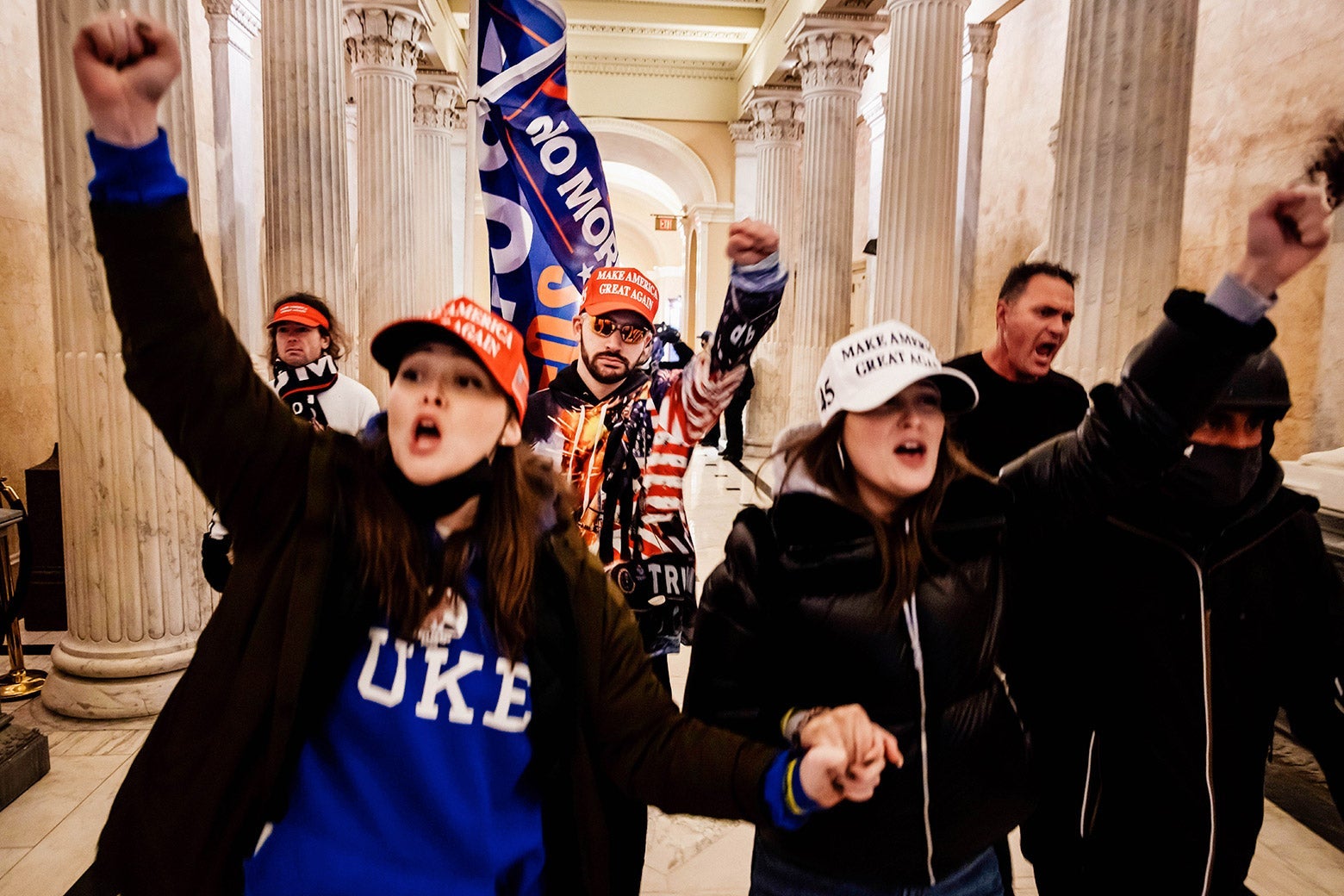 Two women in MAGA hats hold hands and raise their other hands in fists as they walk down a hallway in the Capitol with a small group of people.