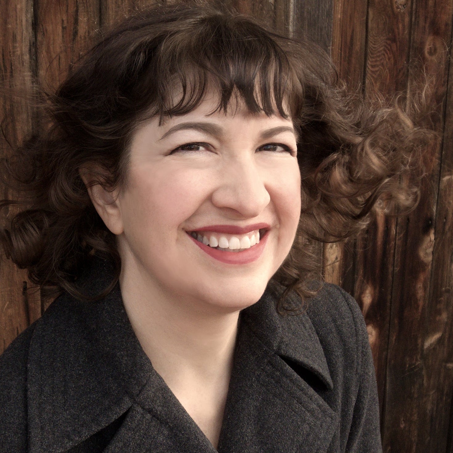 A woman in a gray coat stands in front of a group of trees.
